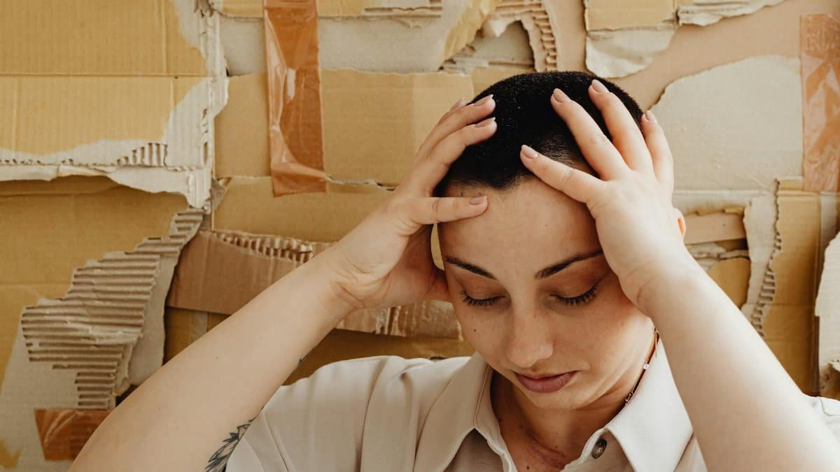 A contemplative woman with a buzz cut rests her hands on her head against a textured cardboard background.