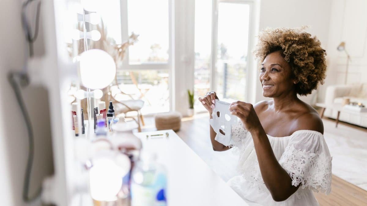 Smiling woman holding face mask in bright, airy room near a vanity mirror.