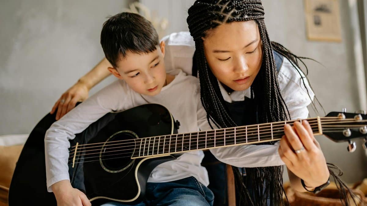 A music teacher instructs a child to play an acoustic guitar indoors.