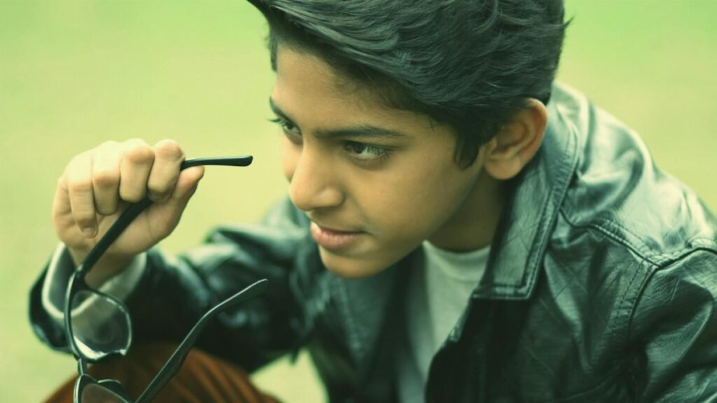 A stylish teenager wearing a black leather jacket holding eyeglasses while posing outdoors.