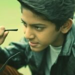 A stylish teenager wearing a black leather jacket holding eyeglasses while posing outdoors.