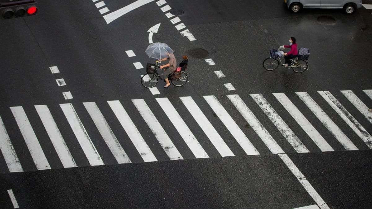 Cyclists navigate a rainy city crosswalk with umbrellas for protection.