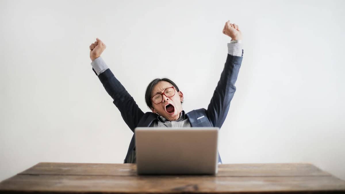 Exhausted man yawning at desk, working on a laptop in an office setting.