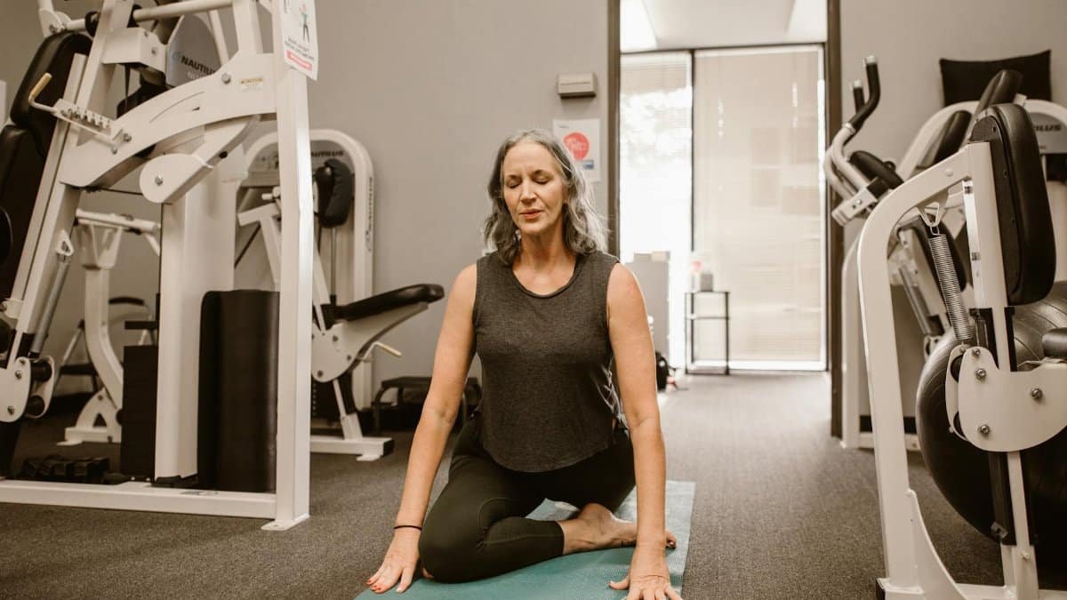 A midlife woman meditates on a yoga mat in a gym, practicing mindfulness and wellness.