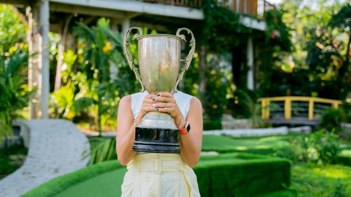 Female golfer holding a large trophy in a lush outdoor setting, celebrating her victory.