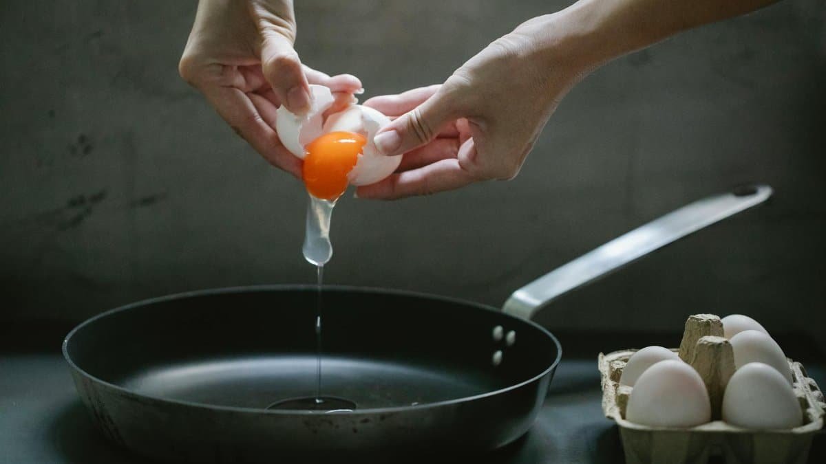Crop anonymous female breaking egg in pan placed on table while cooking breakfast