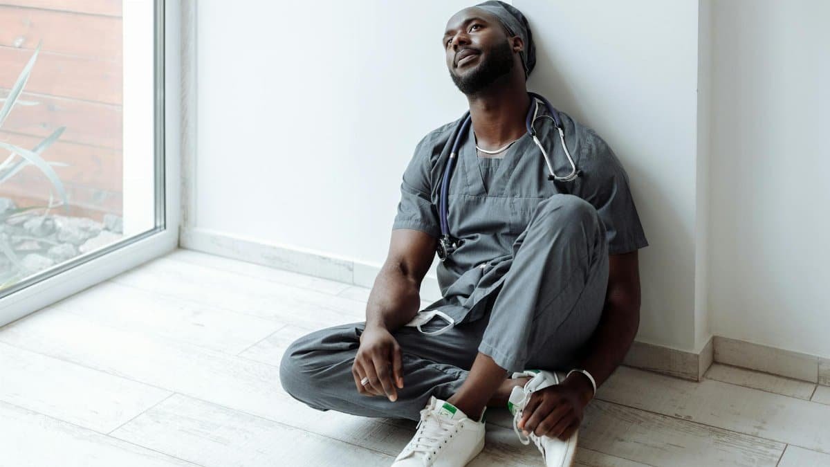 A tired healthcare professional in gray scrubs resting against a wall, reflecting exhaustion.