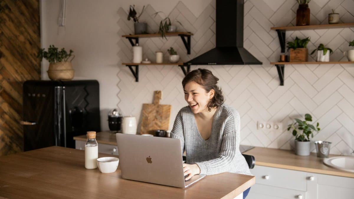 Young female in gray sweater laughing while sitting at wooden table with laptop and milk bottle and having rest watching video during free time