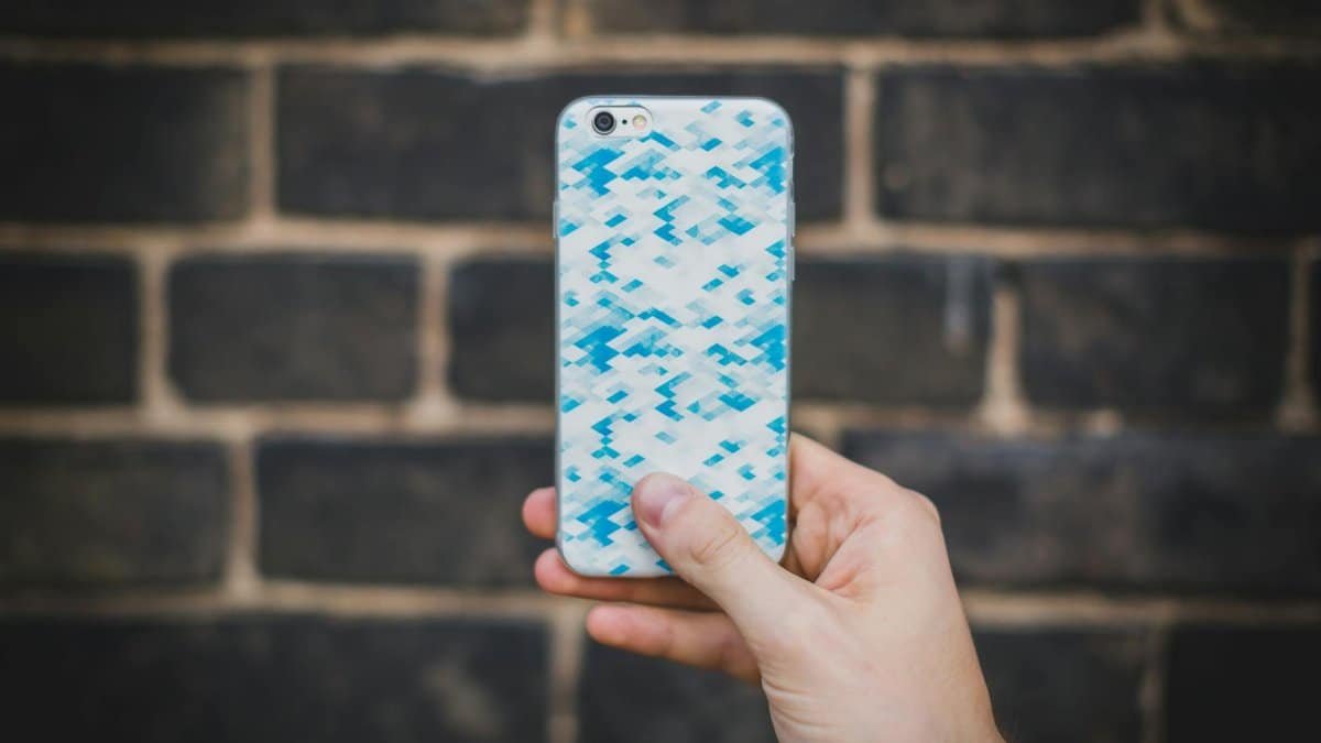 A hand holds a smartphone with a blue patterned case in front of a textured brick wall.