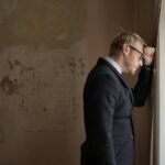 A worried businessman in a suit leaning against a window, expressing stress and concern.