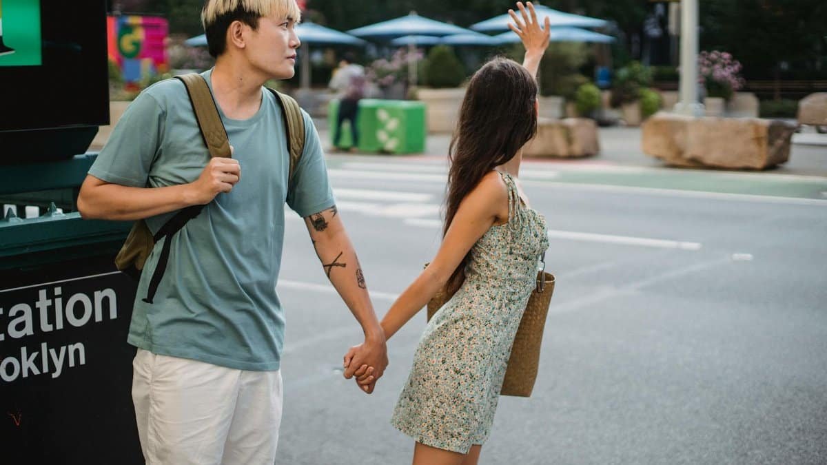 Young female waving hand while catching taxi standing on sidewalk with Asian boyfriend in modern city