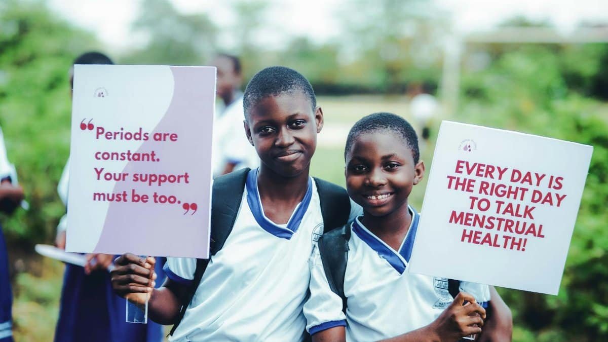 Two students hold signs promoting menstrual health awareness, smiling outdoors.