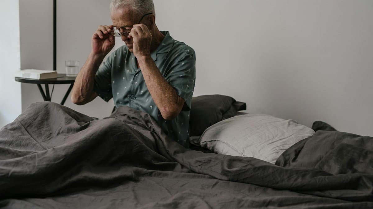 Senior man adjusts glasses while sitting in bed, expressing morning routine and daily life.