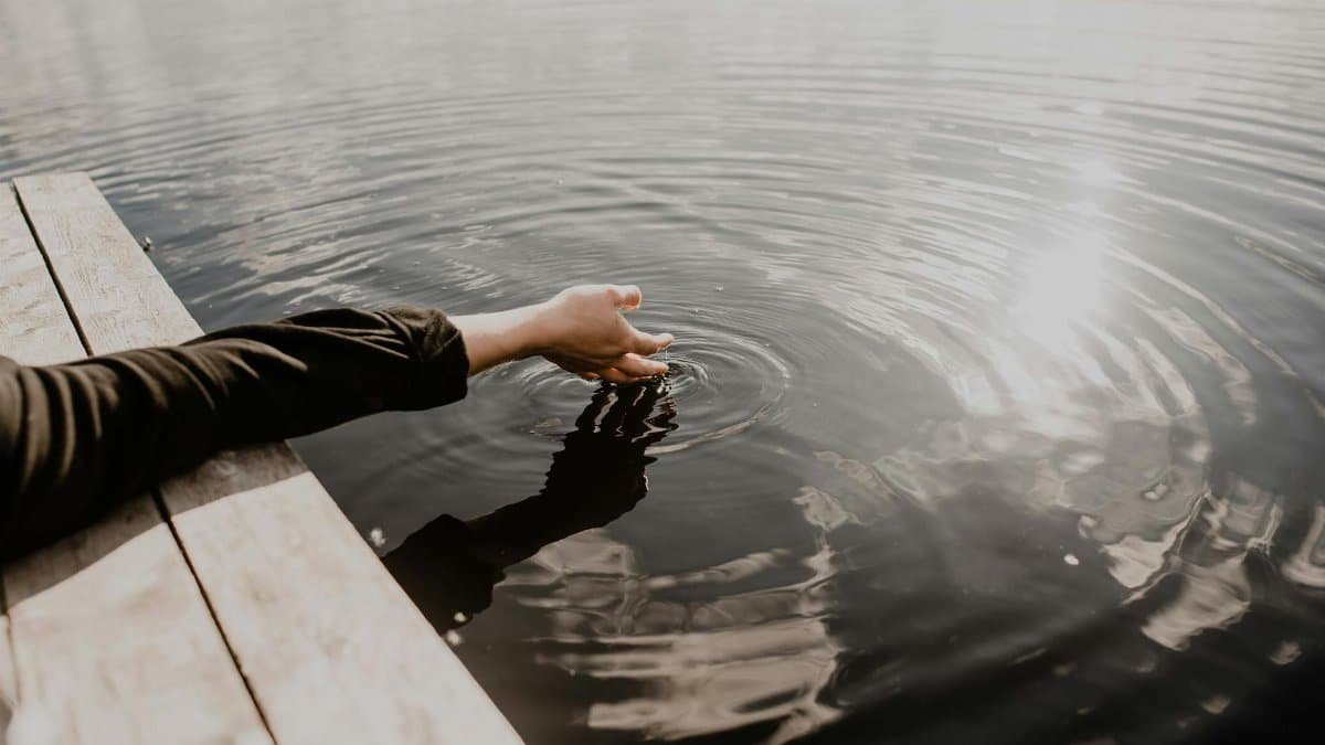 A serene scene of a hand gently touching the calm water surface, creating ripples in the sunlight.