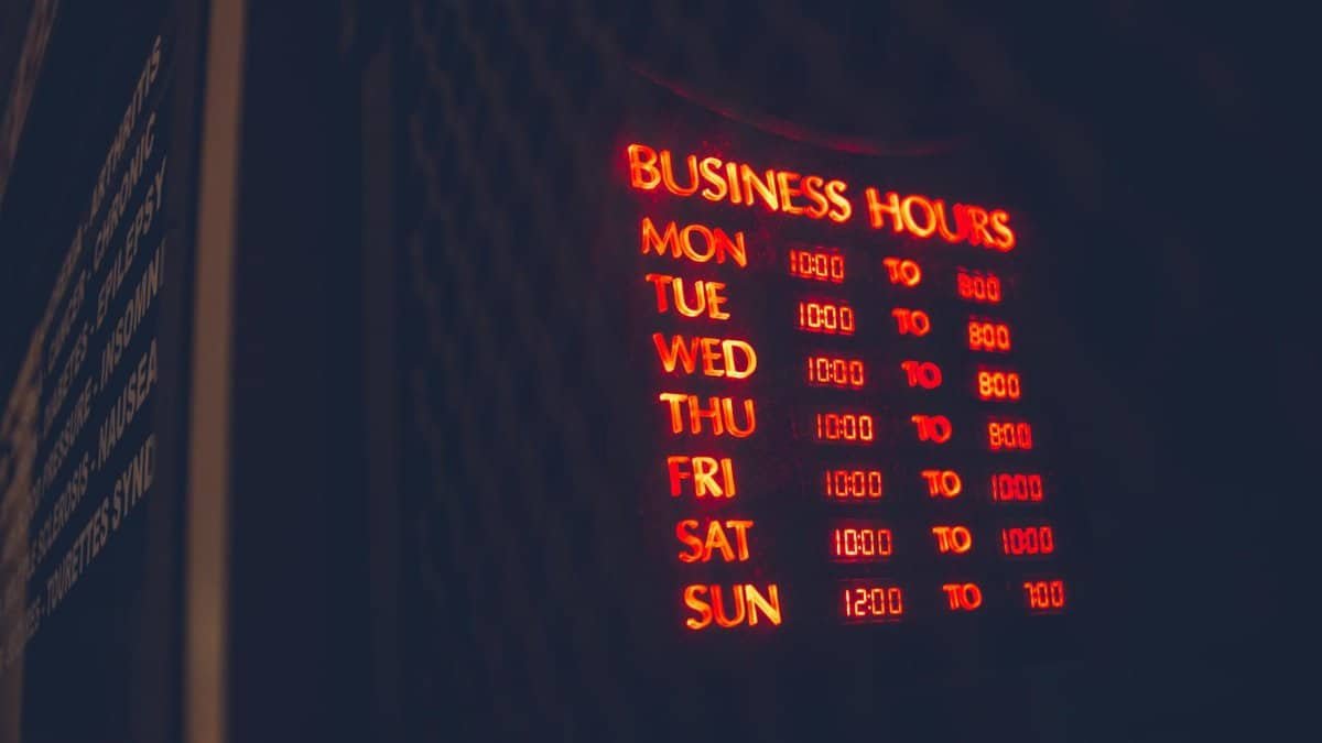 Bright neon sign displaying business hours on a dimly lit wall.