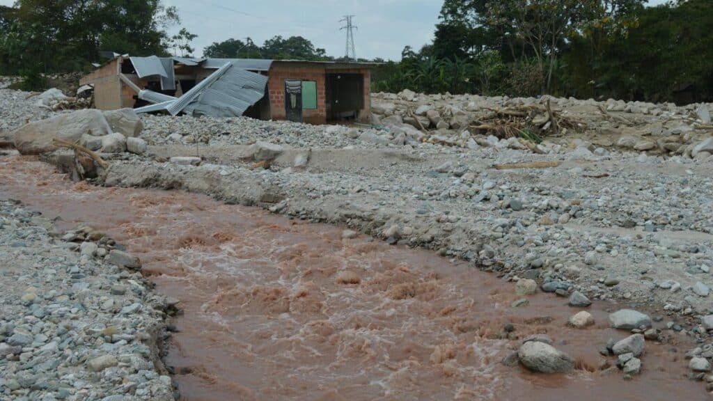 A damaged house stands near a muddy river after a flood, illustrating the impact of natural disasters.
