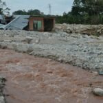 A damaged house stands near a muddy river after a flood, illustrating the impact of natural disasters.