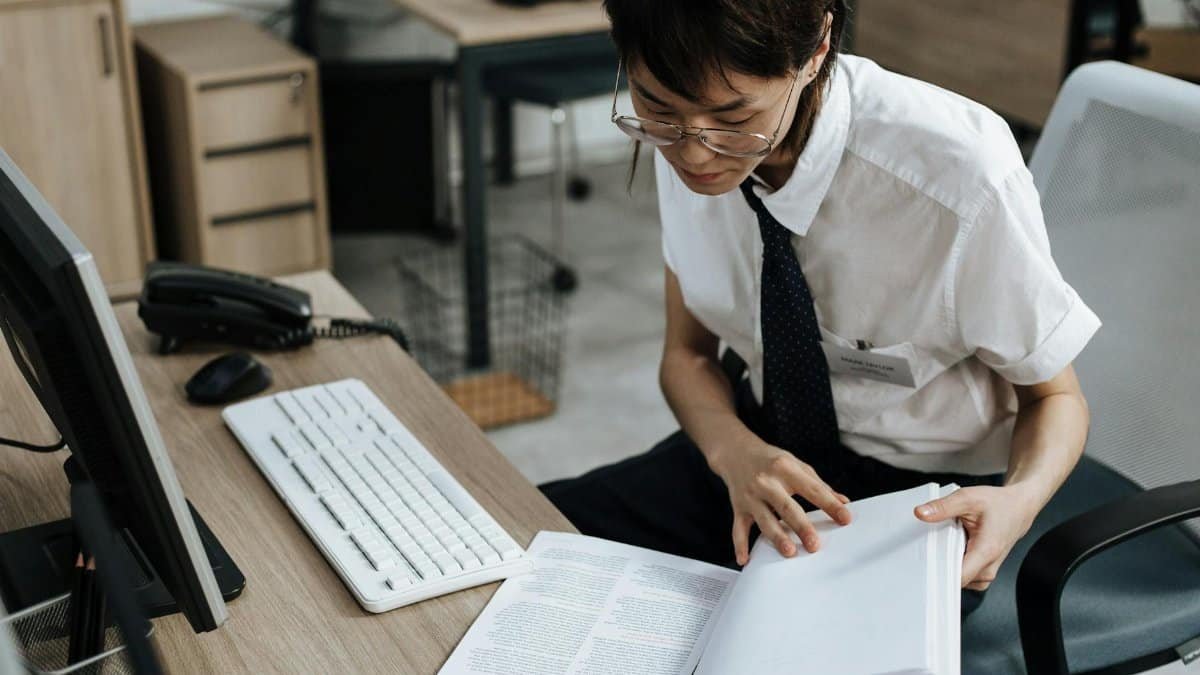 Professional in office attire reviewing documents at a desk with computer and book.