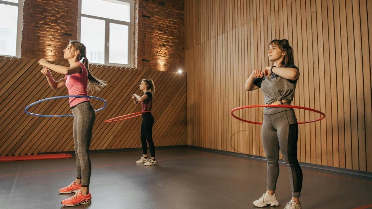Three women exercising with hula hoops in a wooden-paneled gym.