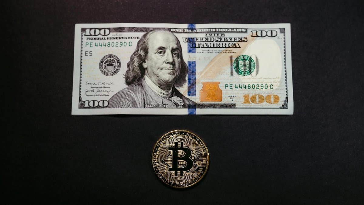 Image of a US 100 dollar bill and a Bitcoin coin on a black background, symbolizing traditional and digital currency.