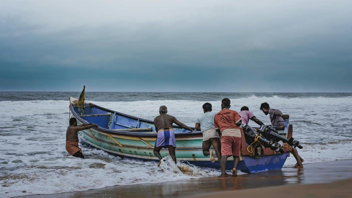 Group of men pushing a fishing boat into the ocean against a cloudy sky, capturing a seaside livelihood moment.