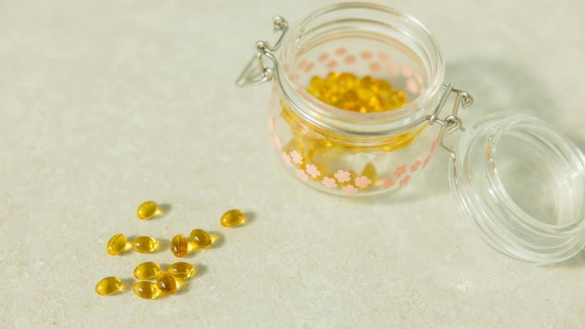 Close-up of open glass jar filled with yellow health capsules on light surface.