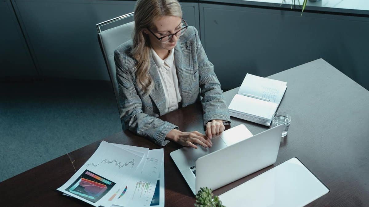 Blonde woman in a blazer working on a laptop in a modern office with documents.