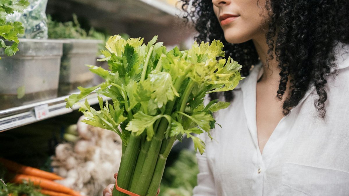 A woman examines a fresh bunch of celery in a produce aisle at the grocery store.