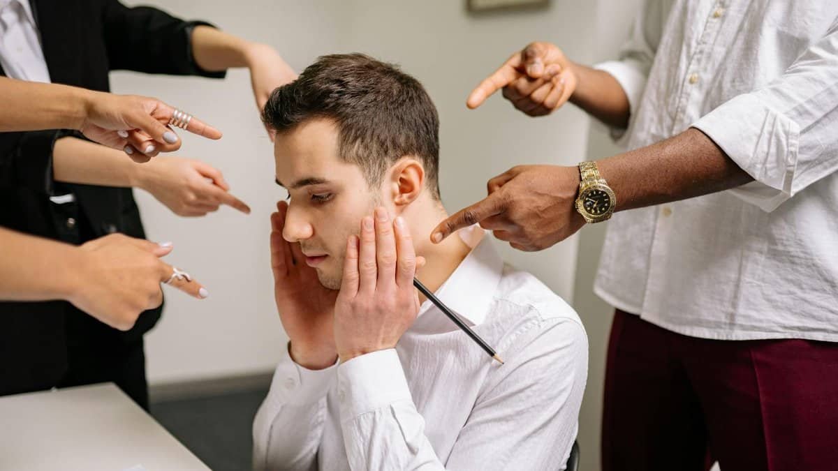 A stressed man at the office surrounded by pointing fingers, representing workplace bullying and stress.