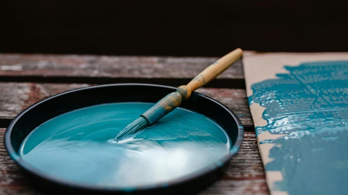 Dirty paintbrush dipped in bowl with azure paint near colorful turquoise carton on wooden bench in street with black background