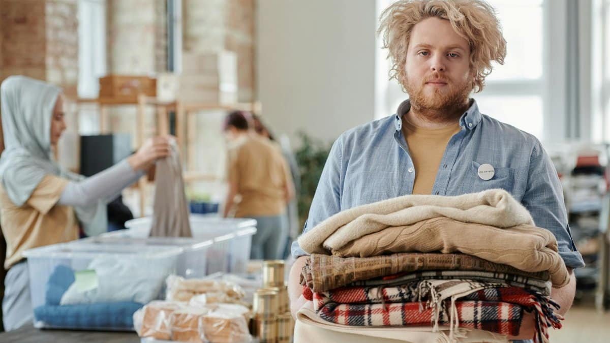 A bearded man volunteers indoors, holding donated clothes for charity work amid a collaborative environment.
