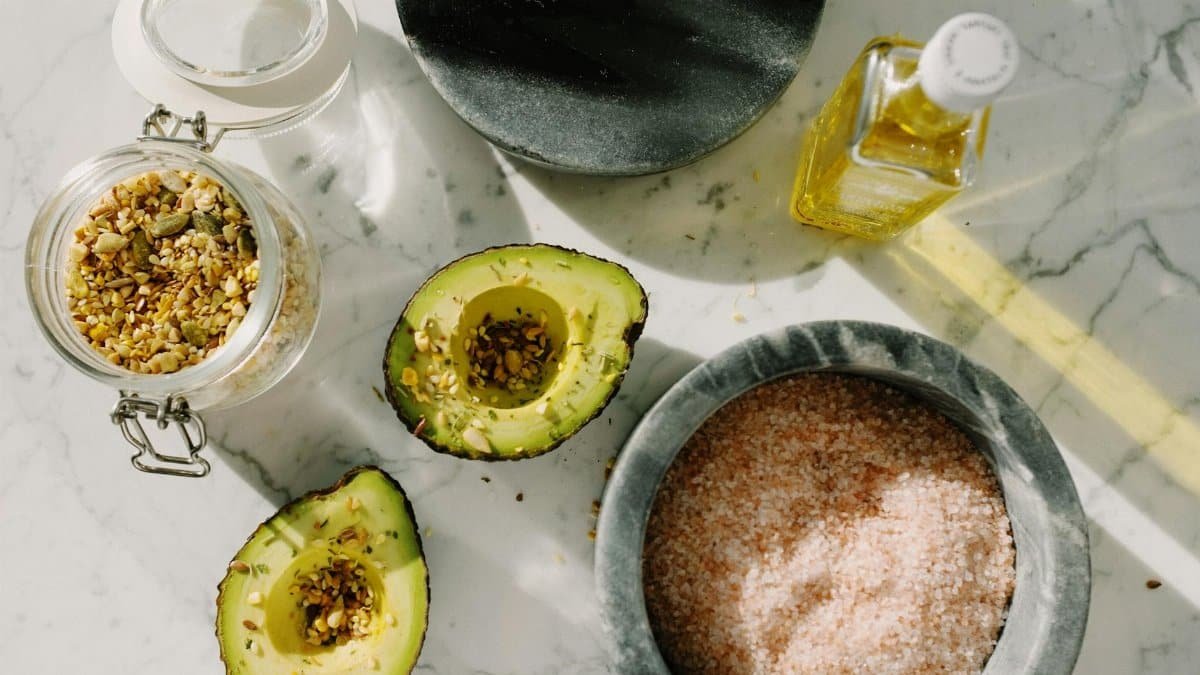 Top view of tasty halved avocado sprinkled with various seeds placed near pink salt with seed mixture in glass pot and other dressing ingredients arranged on white marble table