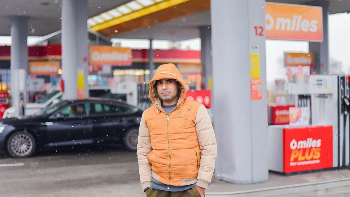 A man in a hooded jacket poses at a snowy gas station, capturing a winter scene.