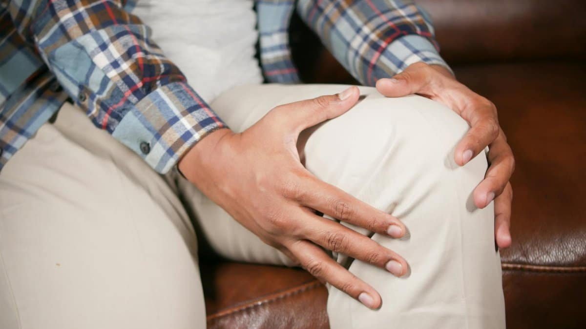 Person sitting on sofa, holding knee in pain. Indoor lifestyle health concept.