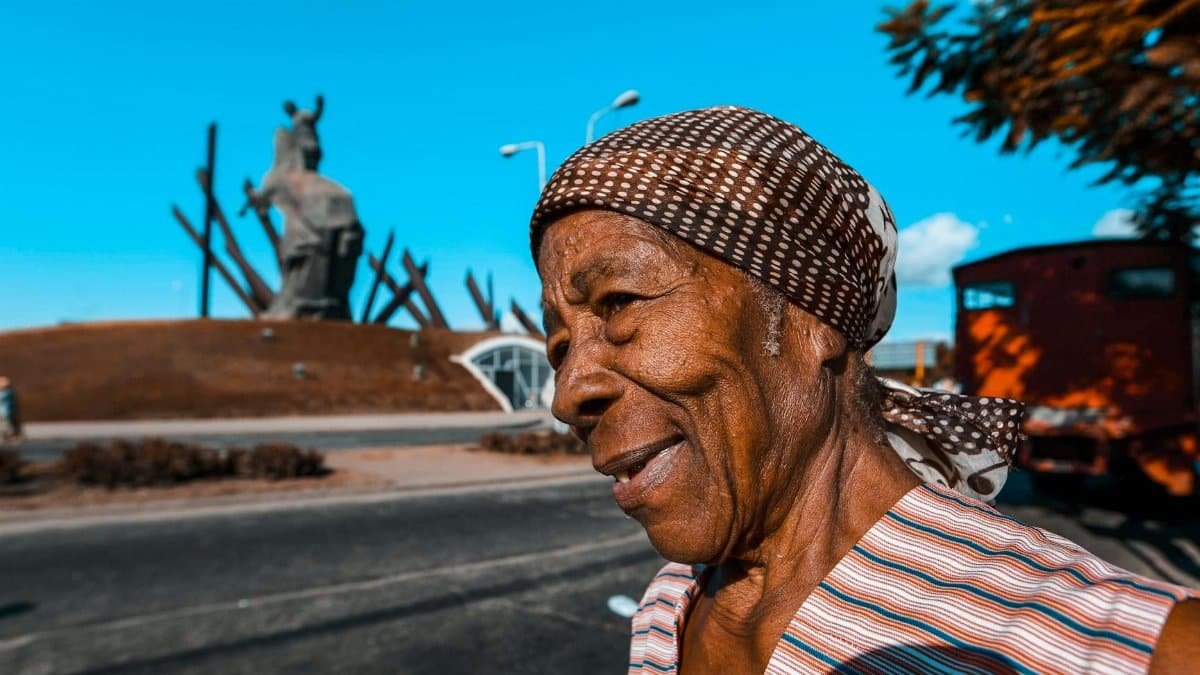 A senior woman in a headscarf smiles outdoors with a statue in the background.