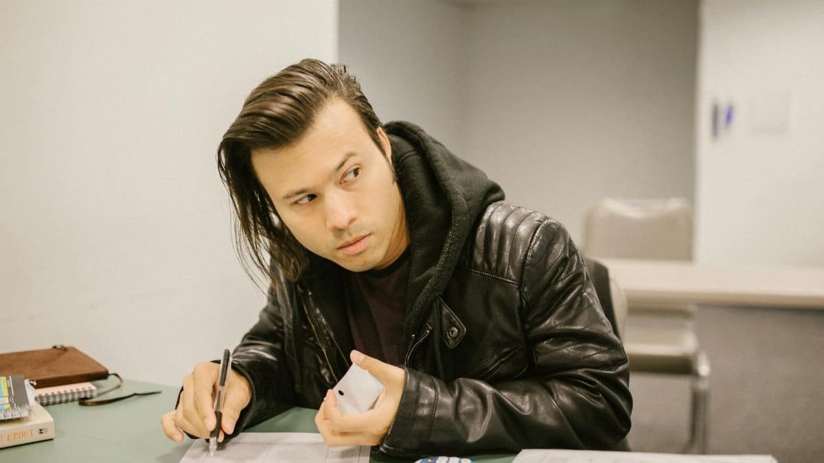 Young man in a classroom looking away while holding a phone, possibly cheating. Exam setting with calculator and paper.