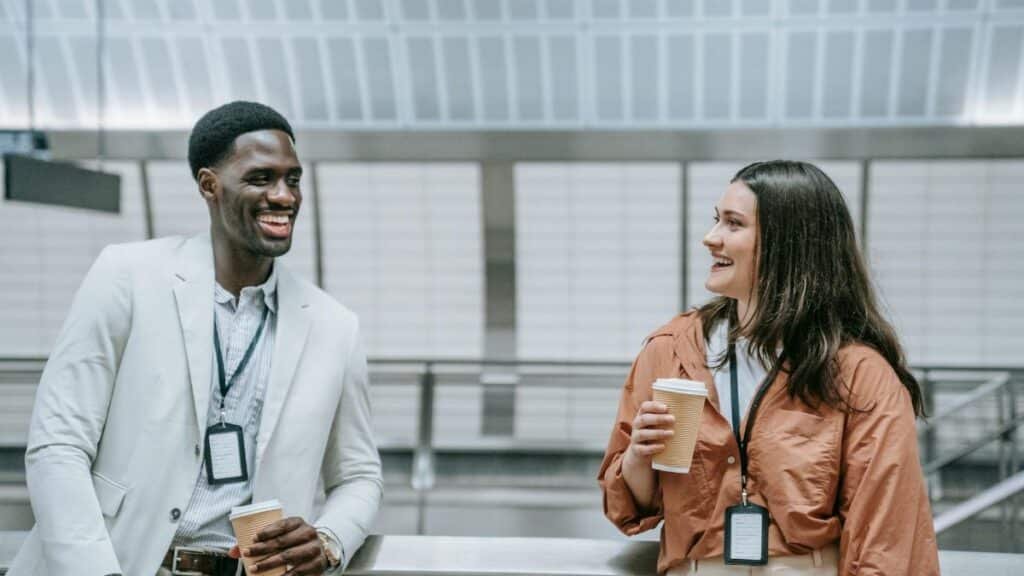 Two smiling coworkers with ID badges enjoy a coffee break indoors.