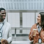 Two smiling coworkers with ID badges enjoy a coffee break indoors.