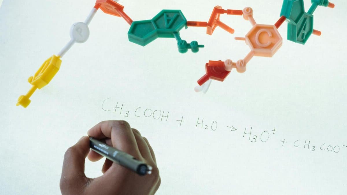 Anonymous chemist writing with marker on illuminated magnetic board formula of Hydronium and Acetate production after studying plastic model of molecule in laboratory