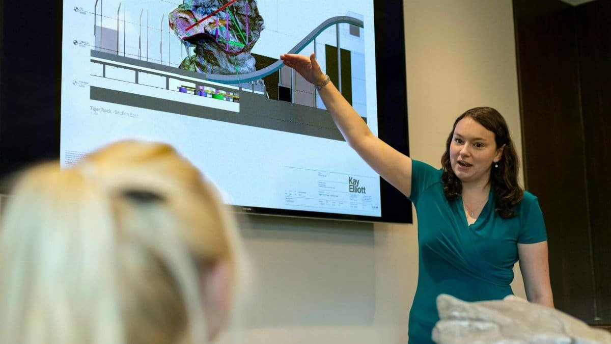 A female engineer delivers a presentation on rollercoaster design using a digital screen in an office.