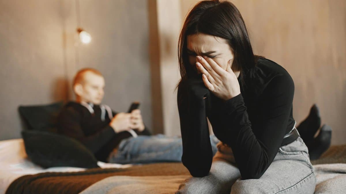 Woman crying on bed, man in background, illustrating relationship stress.