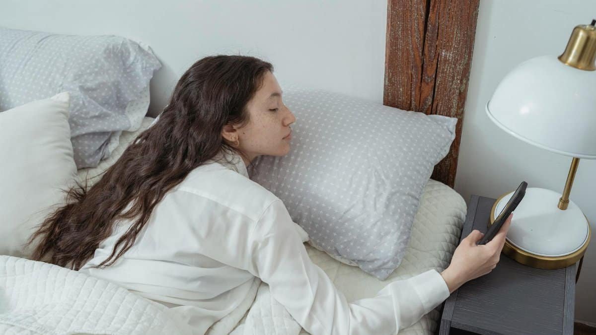 Side view of young brunette with long hair in white nightwear lying on belly in comfortable bed under blanket and using mobile phone in morning at home