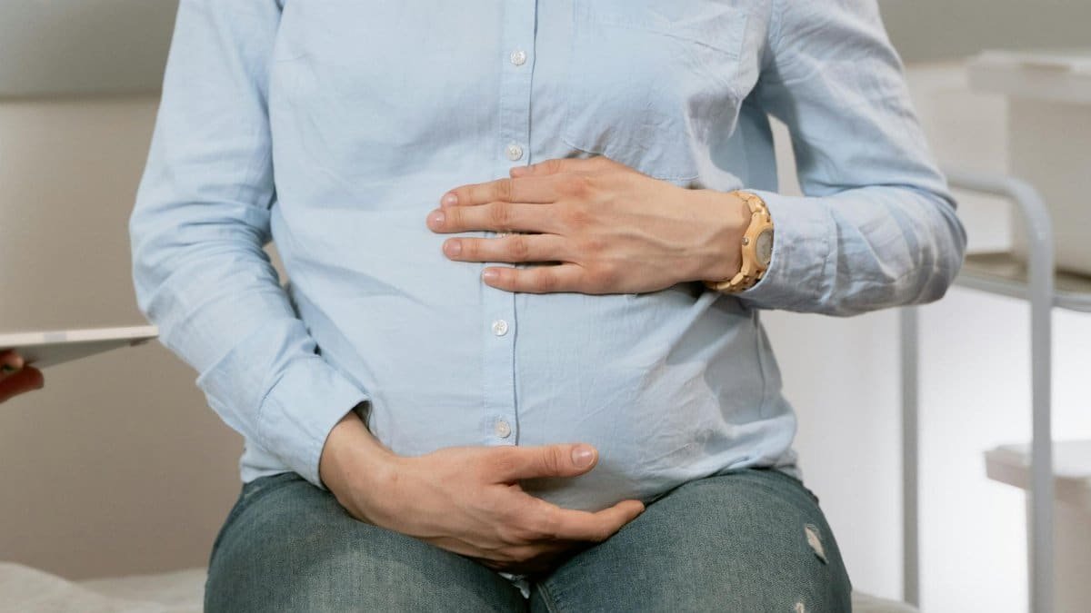 Pregnant woman sitting indoors, hands on belly, wearing casual clothes. Healthcare consultation setting.