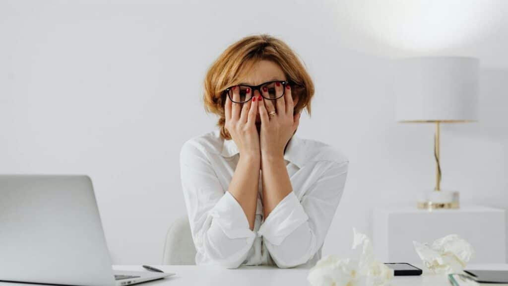 Woman with hands on face, sitting at desk with laptop and tissues, feeling stressed.
