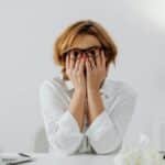 Woman with hands on face, sitting at desk with laptop and tissues, feeling stressed.