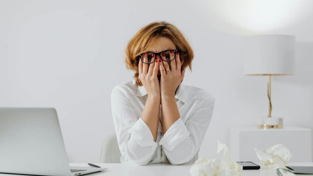 Woman with hands on face, sitting at desk with laptop and tissues, feeling stressed.