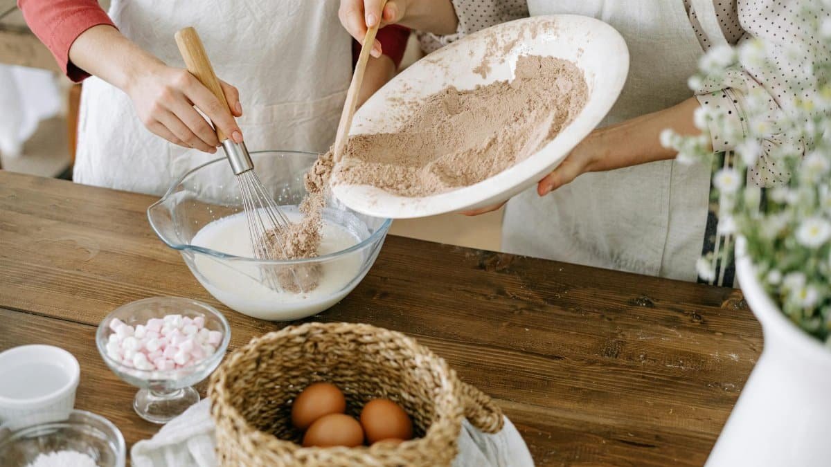 Two women mix ingredients in a rustic kitchen setting, ready for baking.