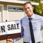 Real estate agent standing with a 'For Sale' sign in front of a house on a sunny day.