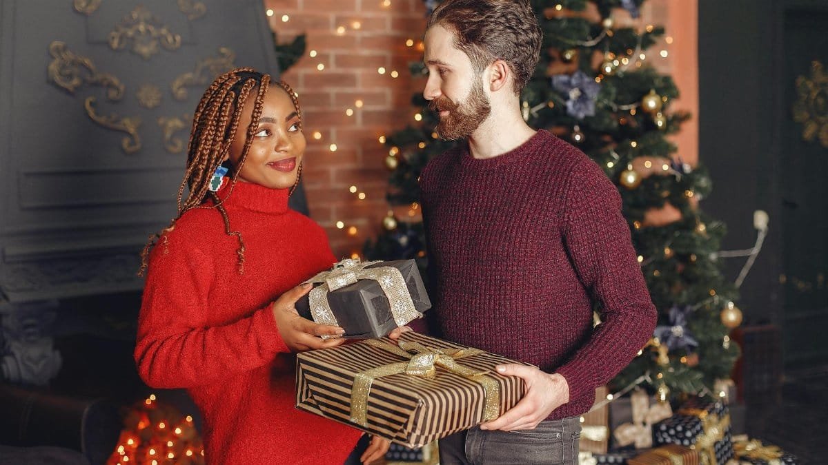 A joyful couple exchanging gifts in front of a beautifully decorated Christmas tree indoors.