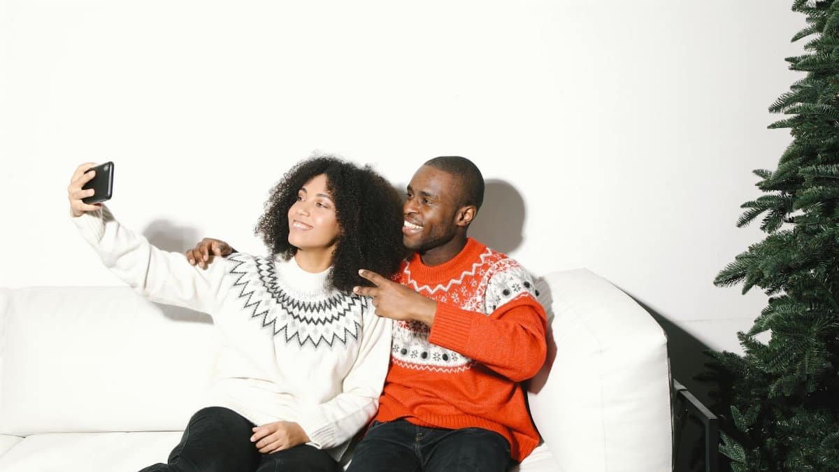 Smiling couple in festive sweaters taking a selfie at home with a Christmas tree.